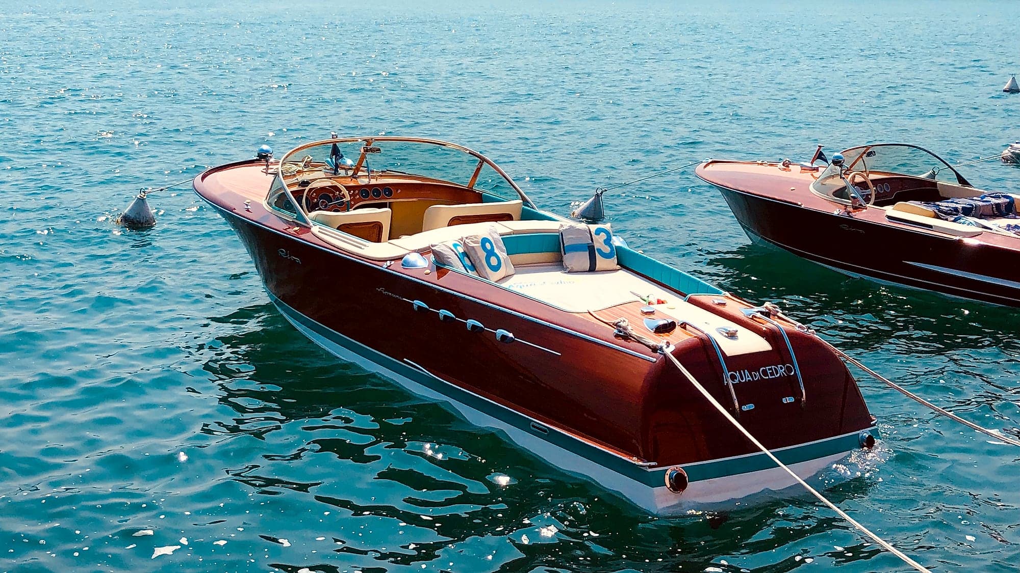 Un motoscafo Riva Aquarama in mogano lucido con i sedili color crema e due cuscini con i numeri 8 e 3, ormeggiato sull'acqua azzurra del lago di Garda, con un secondo motoscafo simile sulla destra