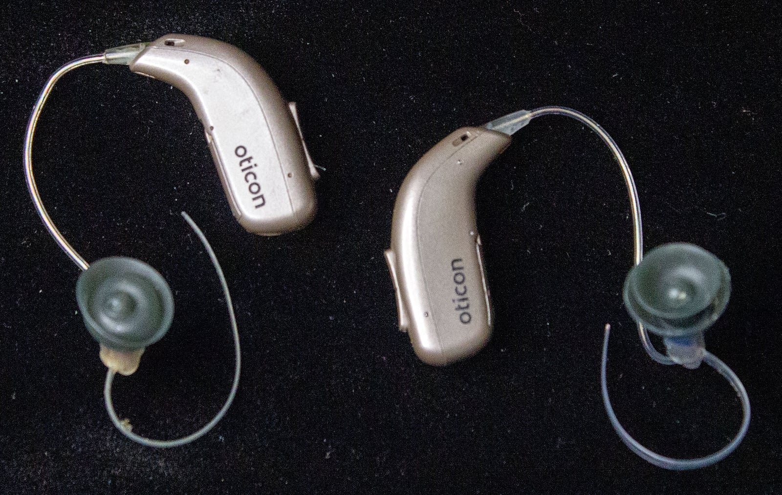 Due apparecchi acustici retroauricolari color champagne con la scritta oticon, ciascuno con il tubicino e la cupola in silicone, appoggiati su un fondo nero