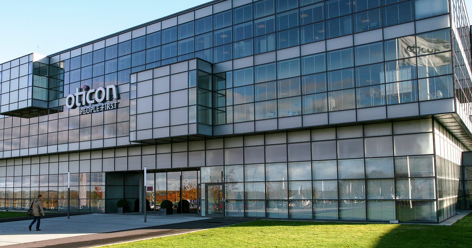 Un grande edificio per uffici con la facciata di vetro e pannelli grigi, la scritta oticon People First sul fianco e un prato davanti all'ingresso
