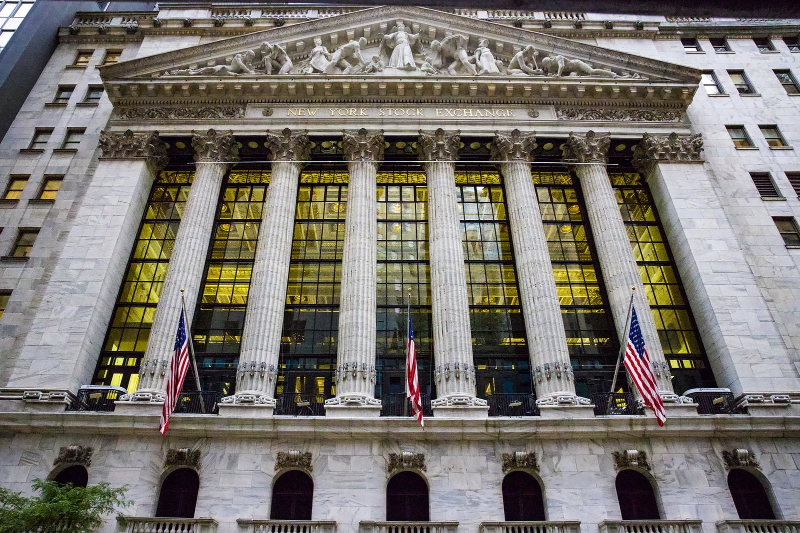 La facciata del New York Stock Exchange vista dal basso: sei colonne corinzie, le vetrate illuminate dall'interno e tre bandiere americane