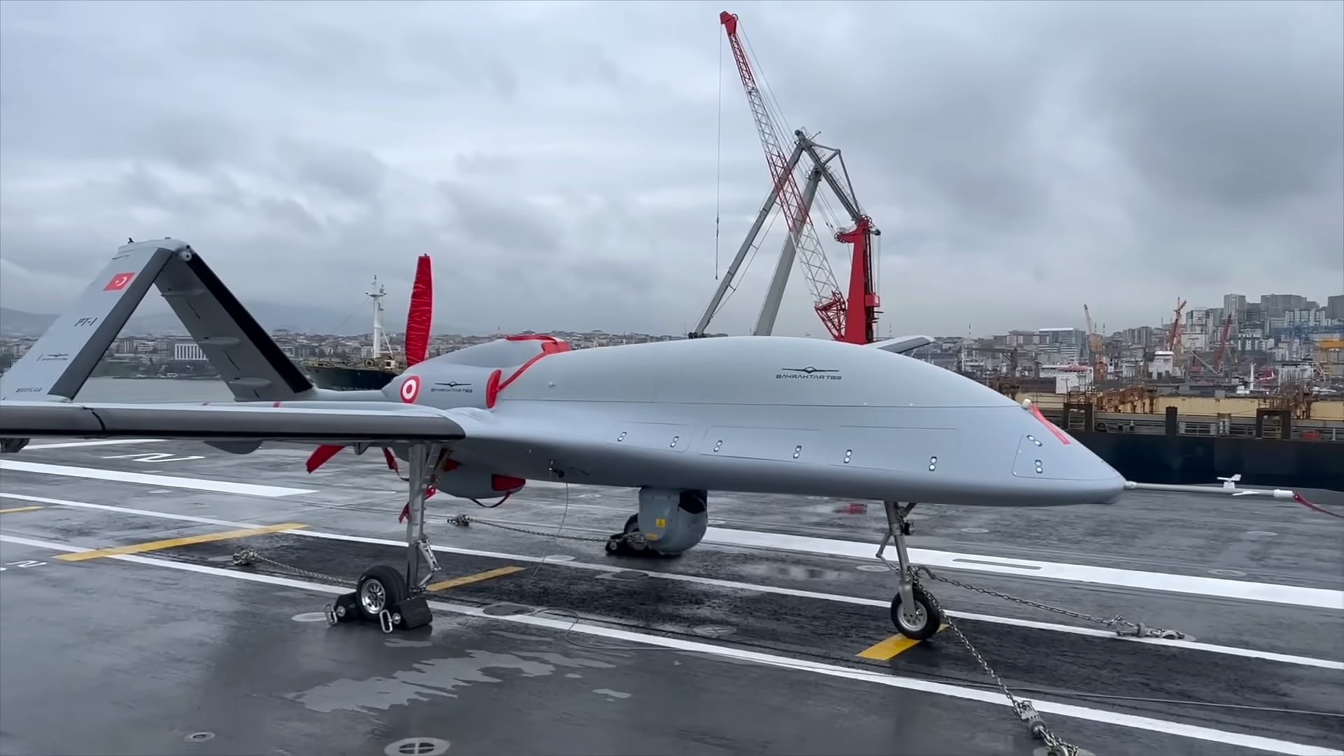 Il drone Bayraktar TB3 sul ponte di volo bagnato di una nave, con le gru di un porto sullo sfondo