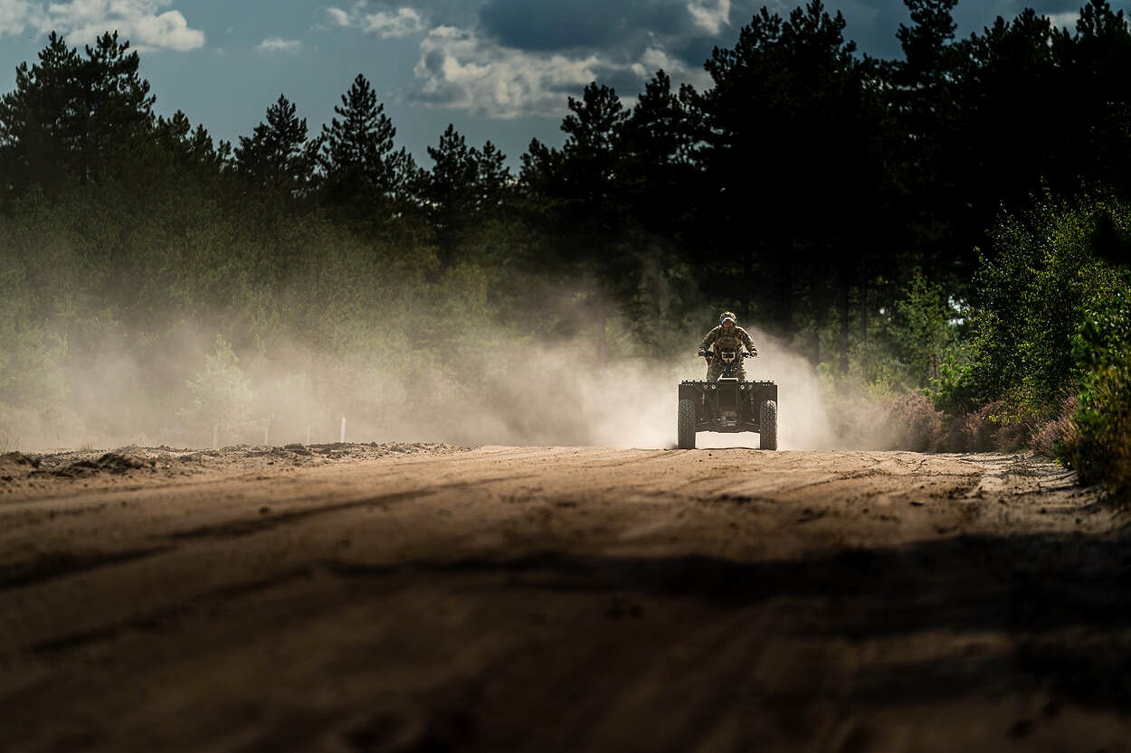 An electric military quad bike travelling alone along a sandy forest track, backlit