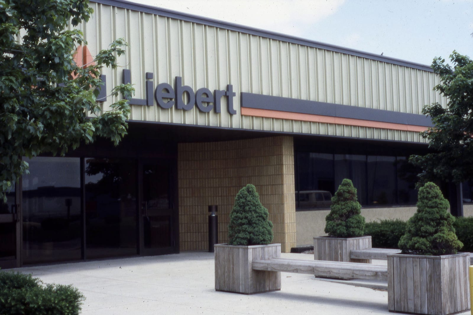 The entrance of a low building with the Liebert sign on its facade and large wooden planters with small fir trees in front of the door